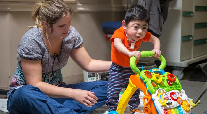 A toddler standing with a push walker beside a therapist